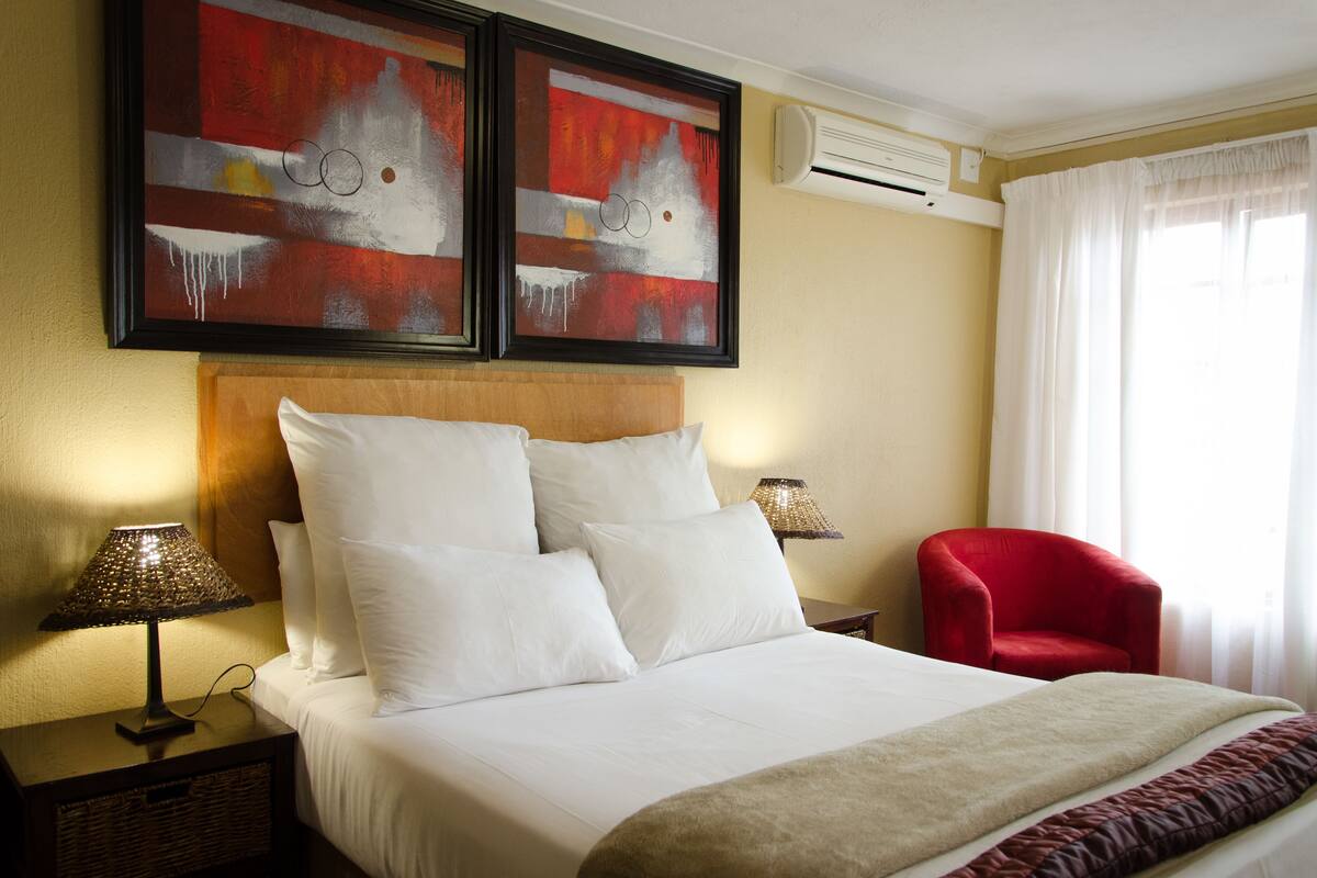 A hotel room with a bed, two paintings, a red chair, and a lamp.
