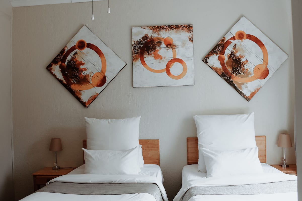 A hotel room with two beds, each with white bedding and wooden headboards. There are three abstract paintings on the wall above the beds.