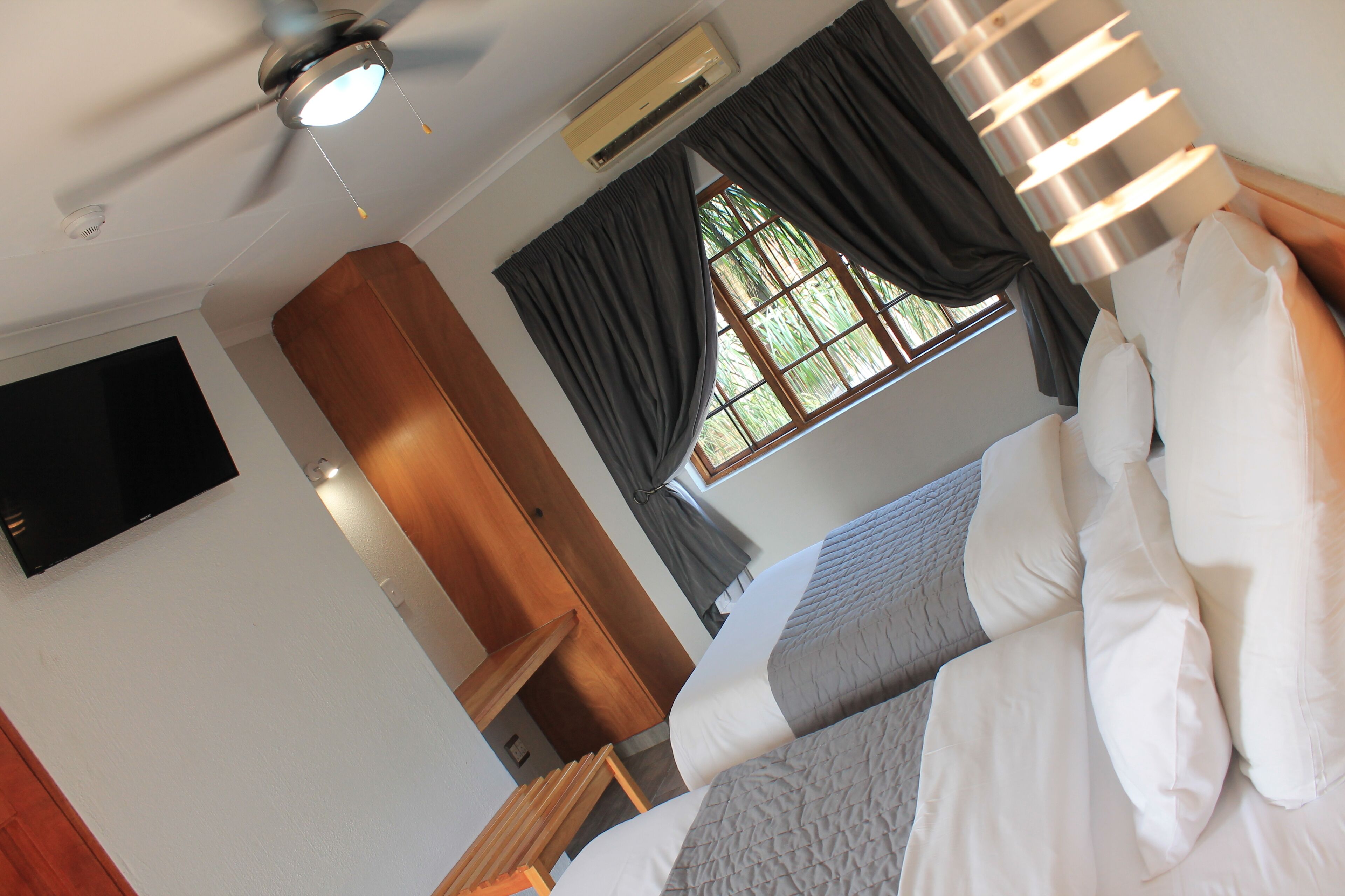 Une chambre d’hôtel équipée d’un lit, d’un ventilateur de plafond, d’un climatiseur, d’un téléviseur fixé au mur et d’une fenêtre avec des rideaux.
