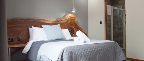 A modern bedroom with a wooden headboard, a grey quilted bedspread, and a glass shower enclosure.