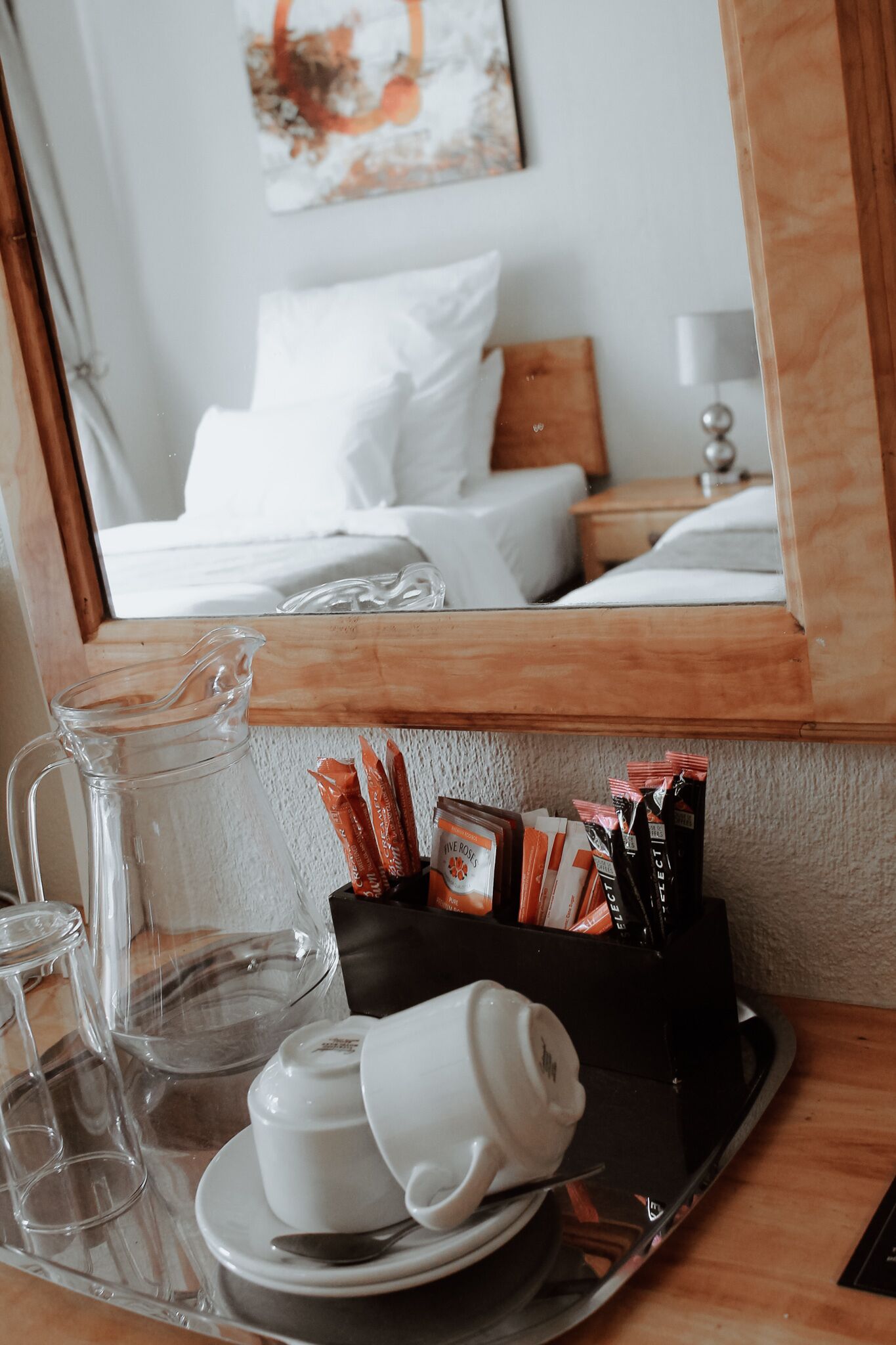 A hotel room with a bed, a bedside table with a tray containing a teapot, cups, and tea bags, and a mirror reflecting the room.