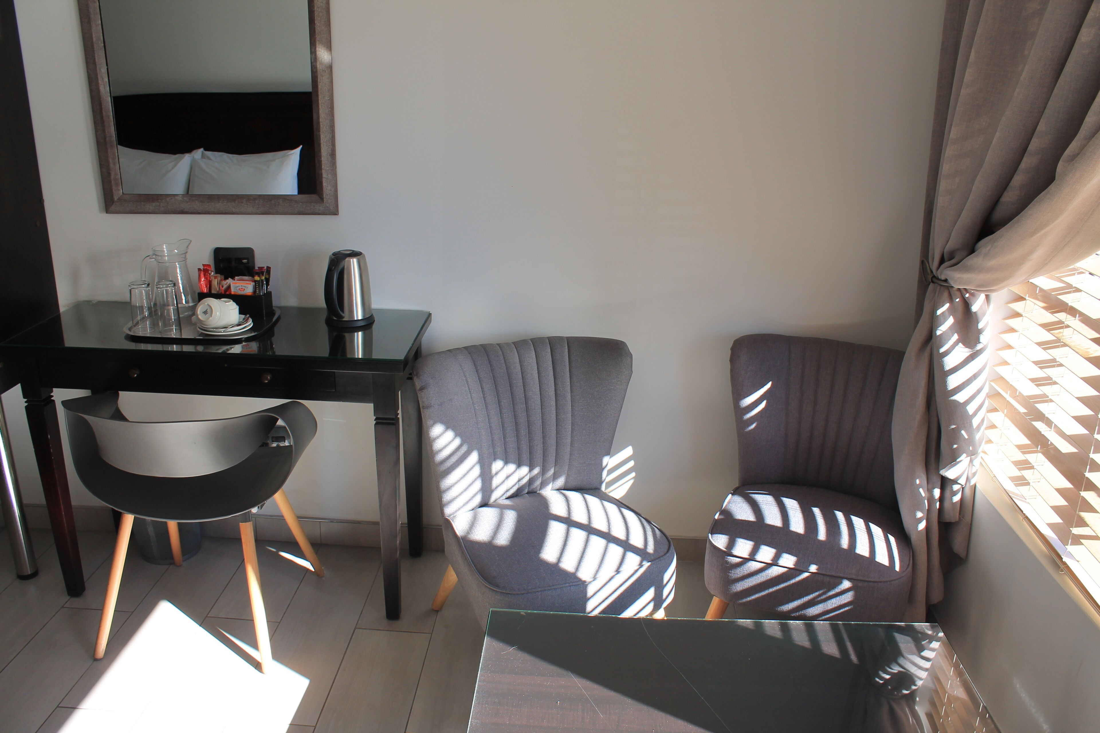 Quarto de hotel moderno com escrivaninha preta, duas cadeiras cinza com padrão, uma mesa pequena e um espelho na parede.