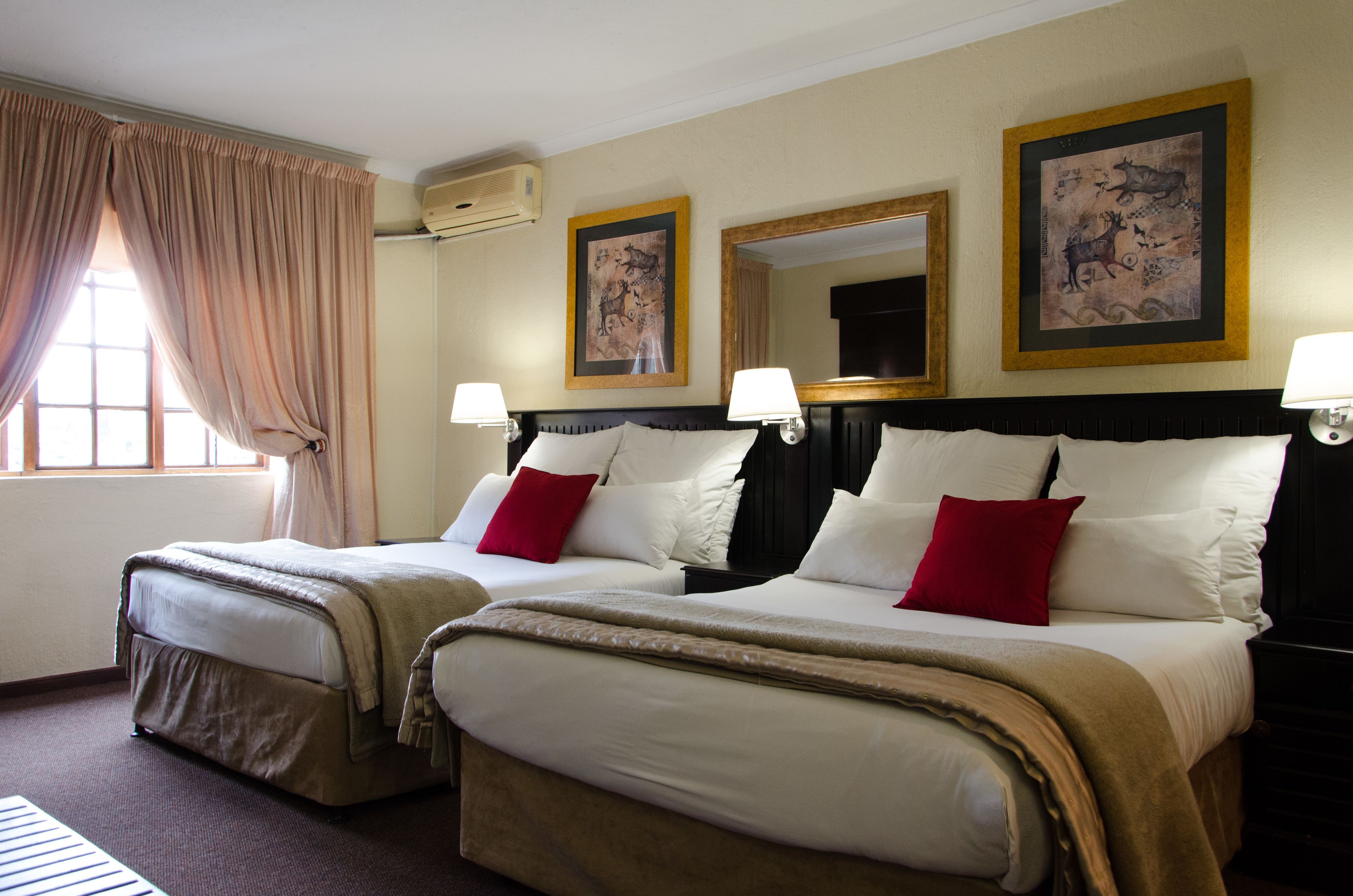 Habitación de hotel con dos camas, cortinas color beige, lámparas fijadas a la pared y cuadros enmarcados.