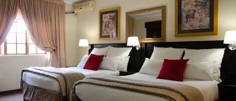 Habitación de hotel con dos camas, cortinas color beige, lámparas fijadas a la pared y cuadros enmarcados.