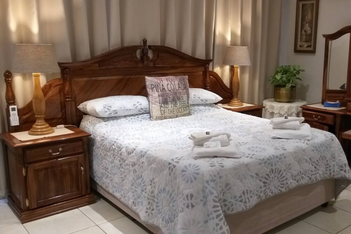 Ein ordentlich bezogenes Bett mit einem blumigen Überwurf, zwei Nachttische mit Lampen, ein kleiner runder Tisch mit einer Pflanze, eine Holzkommode und ein großer Spiegel an der Wand.