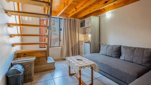 A compact living space with a wooden ceiling, a grey sofa, a small table with a lace tablecloth, a wicker basket, and a compact kitchenette. - Anchorage Guest House (Plettenberg Bay)