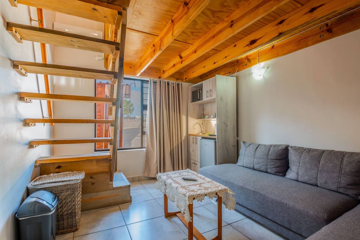Ein gemütlicher Wohnraum mit Holzbalkendecke, einem grauen Sofa, einem kleinen Tisch mit Spitzenunterlage, einem Korbwäschesammler und einer kleinen Küchenzeile.