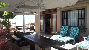 An outdoor seating area with wooden tables and chairs, a covered umbrella, and a view of the street. - Anchorage Guest House (Plettenberg Bay)