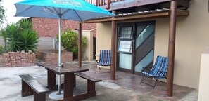 An outdoor patio area with a wooden table and benches, a blue umbrella, and striped chairs. - Anchorage Guest House (Plettenberg Bay)