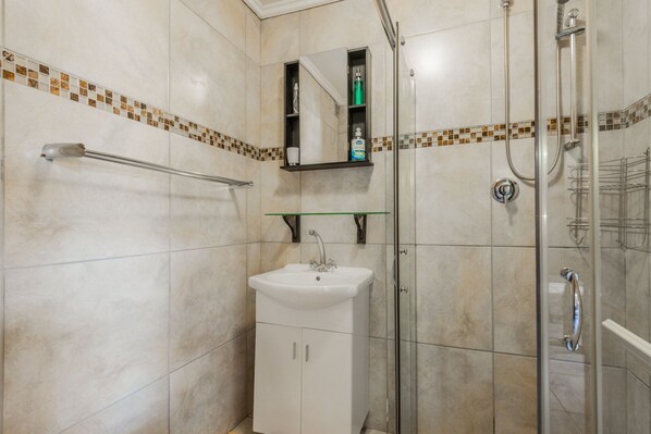 A bathroom with a glass-enclosed shower, a white sink, and a shelf with toiletries. - Anchorage Guest House (Plettenberg Bay)