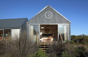 A modern, single-story building with a large glass door leading to a bedroom with a bed, two chairs, and a wooden staircase. - Coot Club (Stanford)
