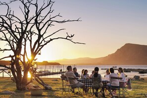 A group of people dining outdoors at sunset with a tree and lanterns in the foreground. - Coot Club (Stanford)