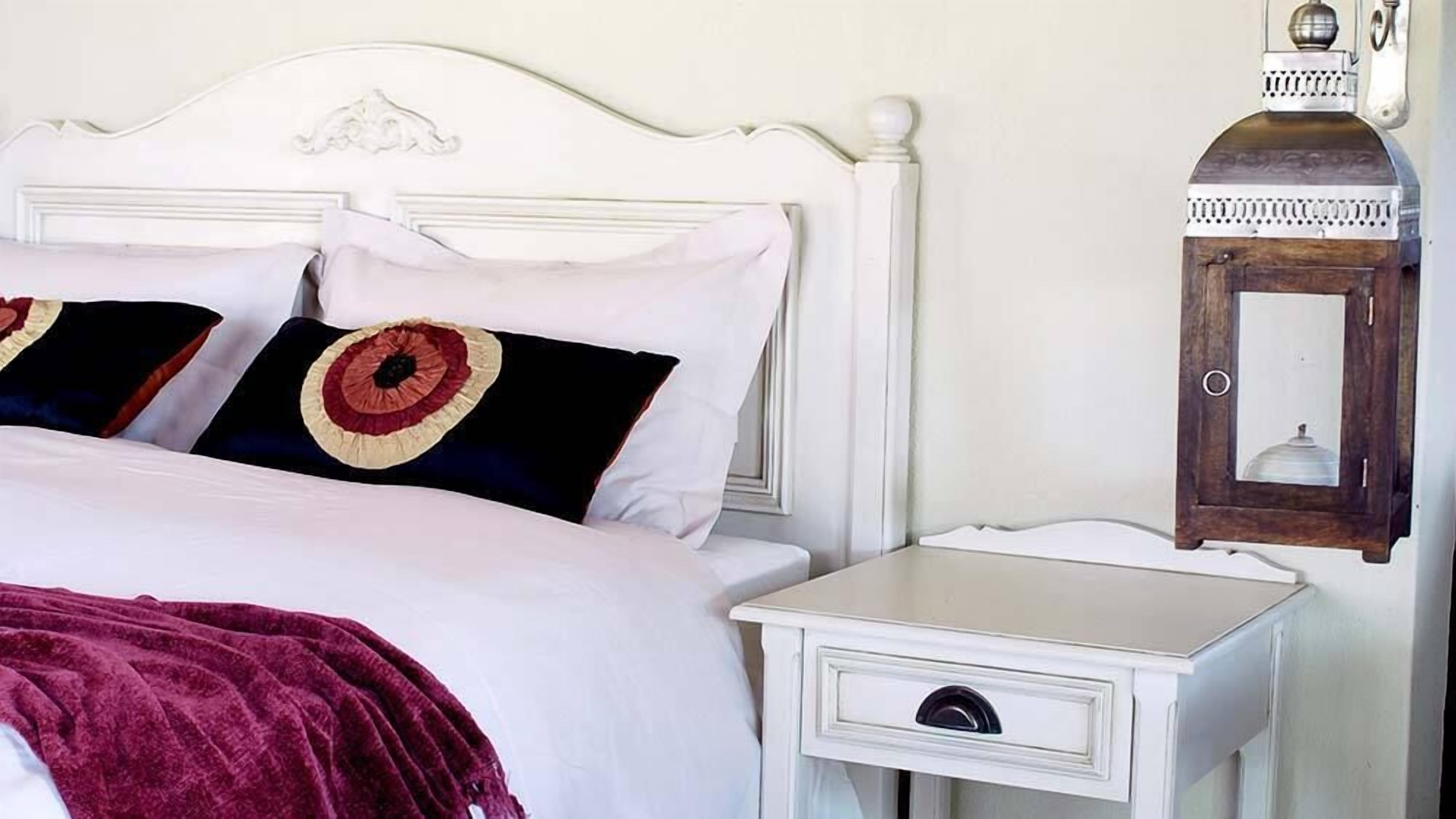 A bed with white bedding and two decorative pillows, a white nightstand with a drawer, and a wall-mounted lantern.