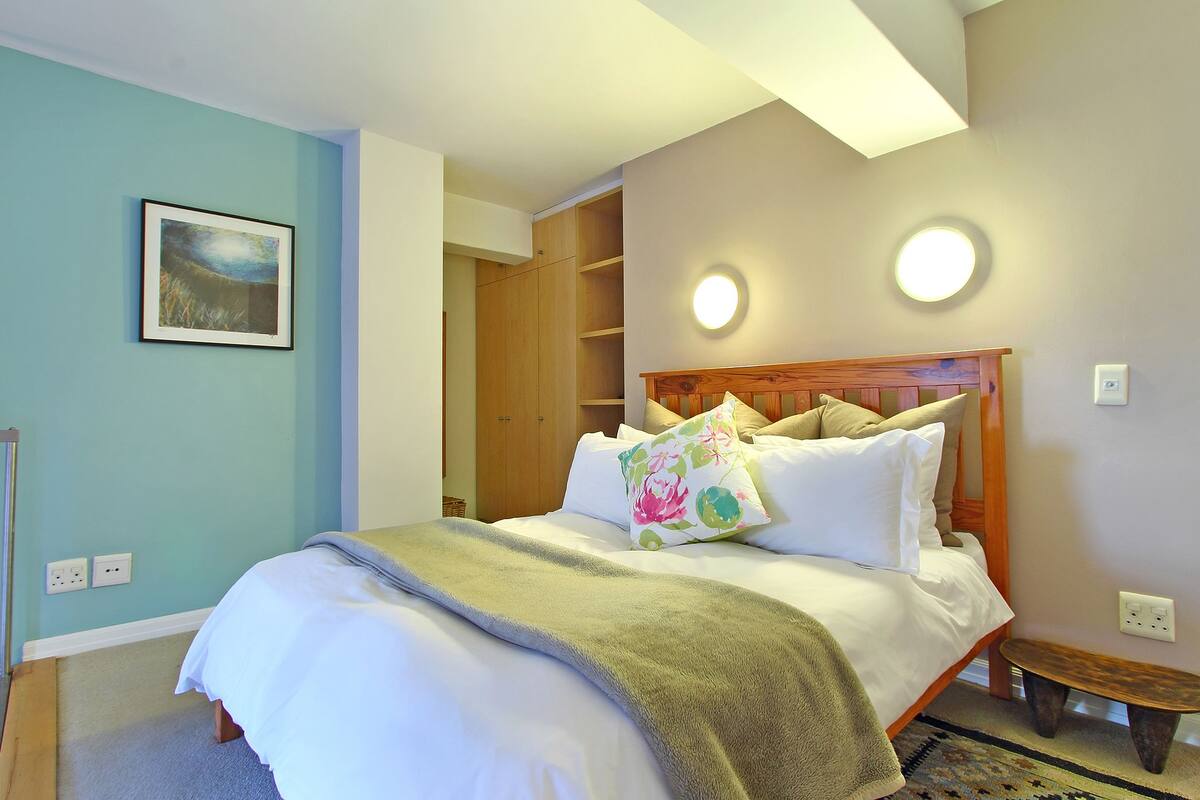 Ein Schlafzimmer mit einem Bett, einem hölzernen Kopfteil, einem Nachttisch und einer Nachttruhe. Über dem Bett befindet sich eine Wandleuchte und an der Wand eine Steckdose.