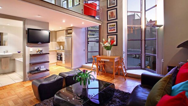 A modern apartment with a kitchen, dining area, and living room. It features a balcony, large windows, and a flat-screen TV. - Cape eazi stayz Old Mutual Apartments (Cape Town)