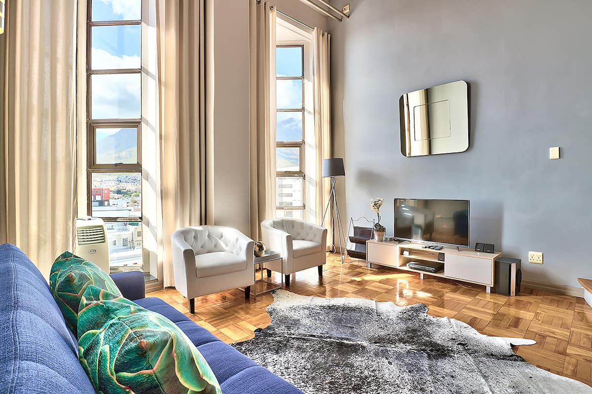 Ein modernes Wohnzimmer mit einer Couch, zwei Sesseln, einem Fernseher und einem großen Fenster mit Vorhängen.
