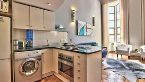 A compact kitchen with a built-in washer and dryer, a stove, and a sink. There is a dining area with a blue couch and a white chair. The room has a large window and a view of the city. - Cape eazi stayz Old Mutual Apartments (Cape Town)