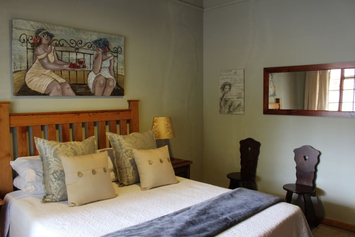 Un dormitorio con cama, almohadas, una mesita de noche, lámpara, espejo y un cuadro en la pared.