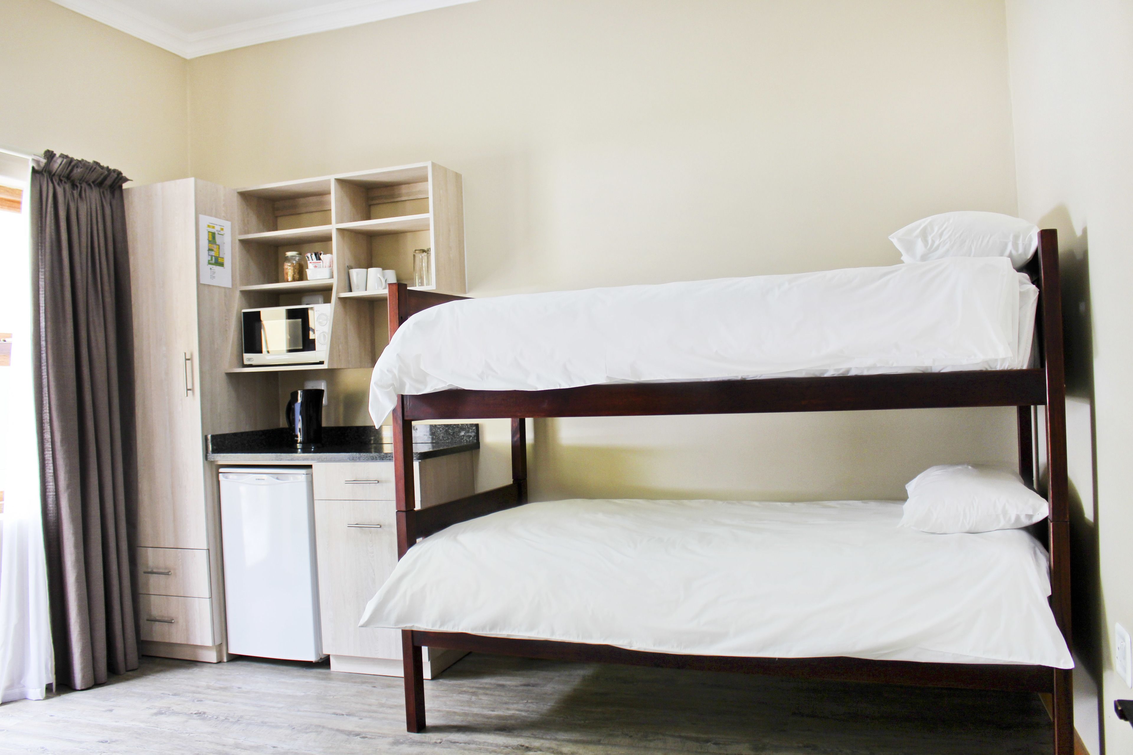 Una cama litera con ropa blanca, una kitchenette con microondas y un armario con estantes.
