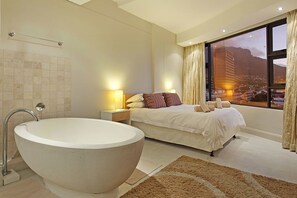 A modern bedroom with a large bathtub, a bed with white bedding, and a view of a cityscape through a large window. - Afribode Acc Piazza (Cape Town)