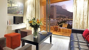 A modern living room with a sofa, ottoman, and a coffee table with a vase of flowers. A flat-screen TV is on a white shelf, and there's a large window with a view of a cityscape and mountains. - Afribode Acc Piazza (Cape Town)