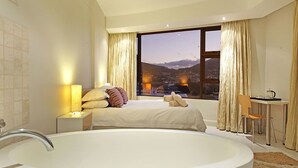 A modern bedroom with a large bathtub, a bed with pillows, a desk, and a view of a cityscape through a large window. - Afribode Acc Piazza (Cape Town)