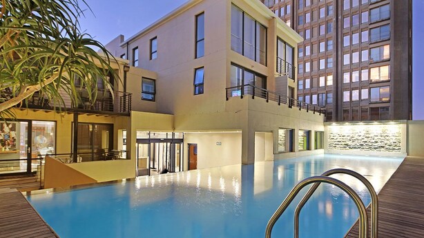 A modern hotel with a large indoor pool, a balcony, and a view of a high-rise building. - Afribode Acc Piazza (Cape Town)