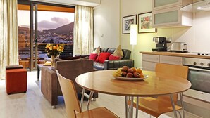 A modern hotel room with a dining area, a sofa with cushions, a coffee machine, and a view of a cityscape. - Afribode Acc Piazza (Cape Town)