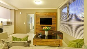 Una sala de estar moderna con una televisión de pantalla plana montada en una pared de madera, una mesa de centro de vidrio con un jarrón de flores y un sofá con cojines verdes.