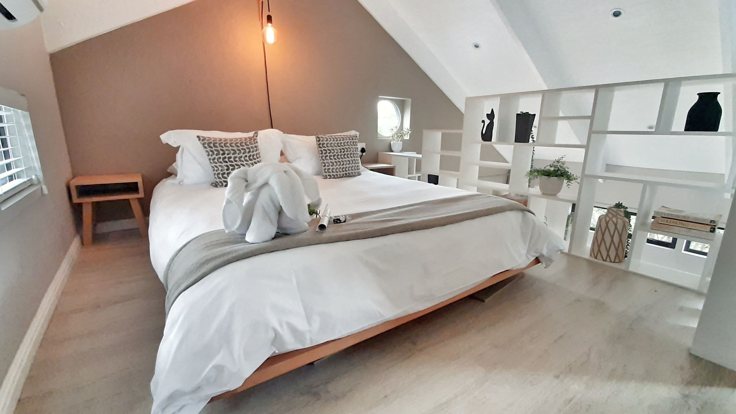 A modern bedroom with a sloped ceiling, a large bed with white bedding, a bedside table, a small stool, and a shelf with decorative items.