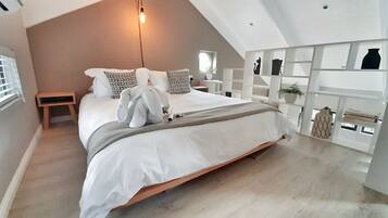 A modern bedroom with a sloped ceiling, a large bed with white bedding, a bedside table, a small stool, and a shelf with decorative items.