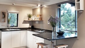 Une cuisine moderne avec des meubles blancs, un plan de travail noir et un îlot central intégré avec des tabourets. Une fenêtre laisse entrer la lumière naturelle et offre une vue sur la verdure à l’extérieur.