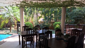 Un área de comedor al aire libre con mesas y sillas de madera, rodeada de abundante vegetación y una piscina.