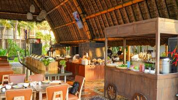 An outdoor dining area with a thatched roof, wooden tables, and chairs. There are food and drink stations with various items displayed.