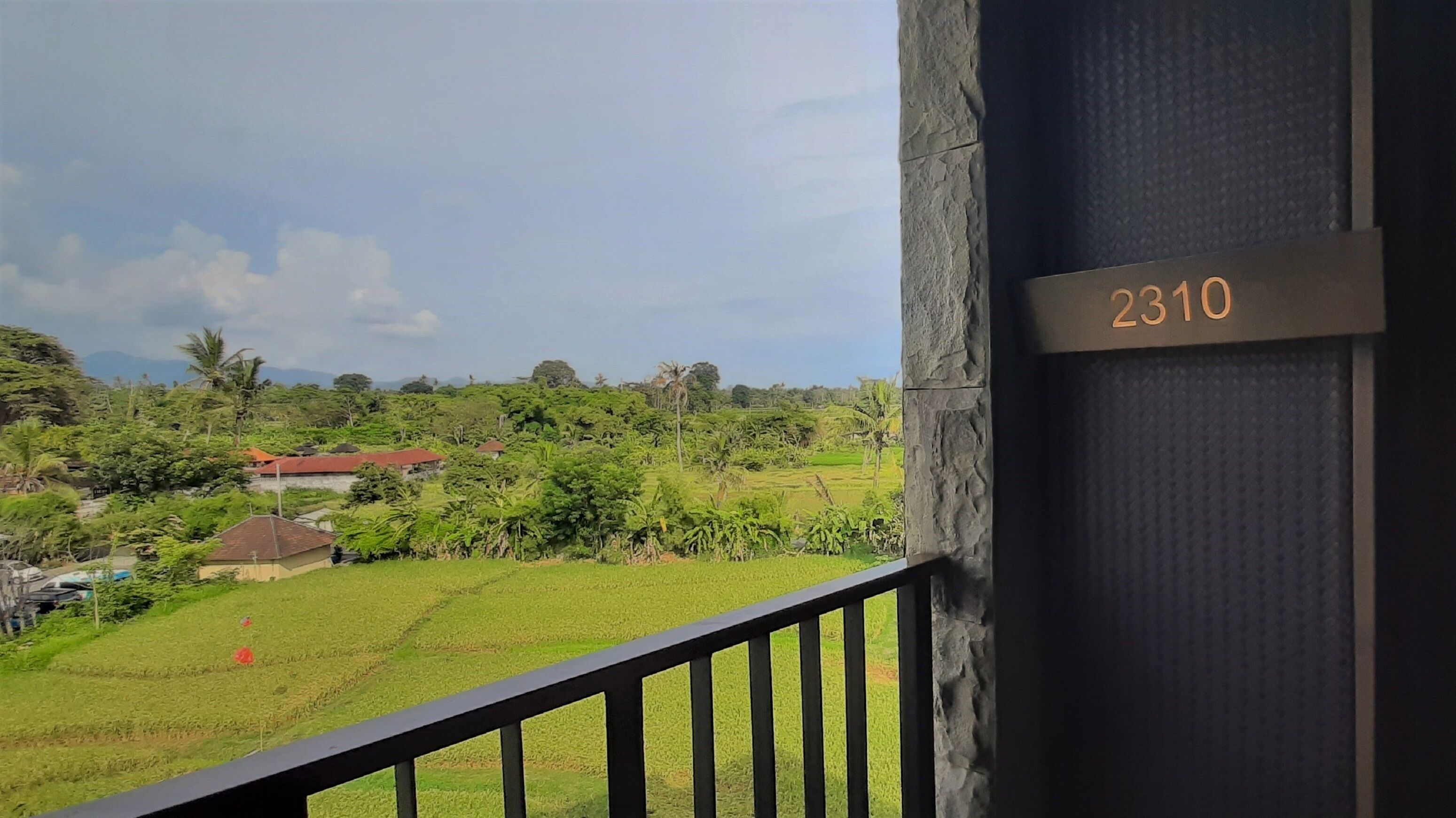 A balcony with a view of a lush green landscape, including fields and a distant building. A sign with the number 2310 is visible on the wall.