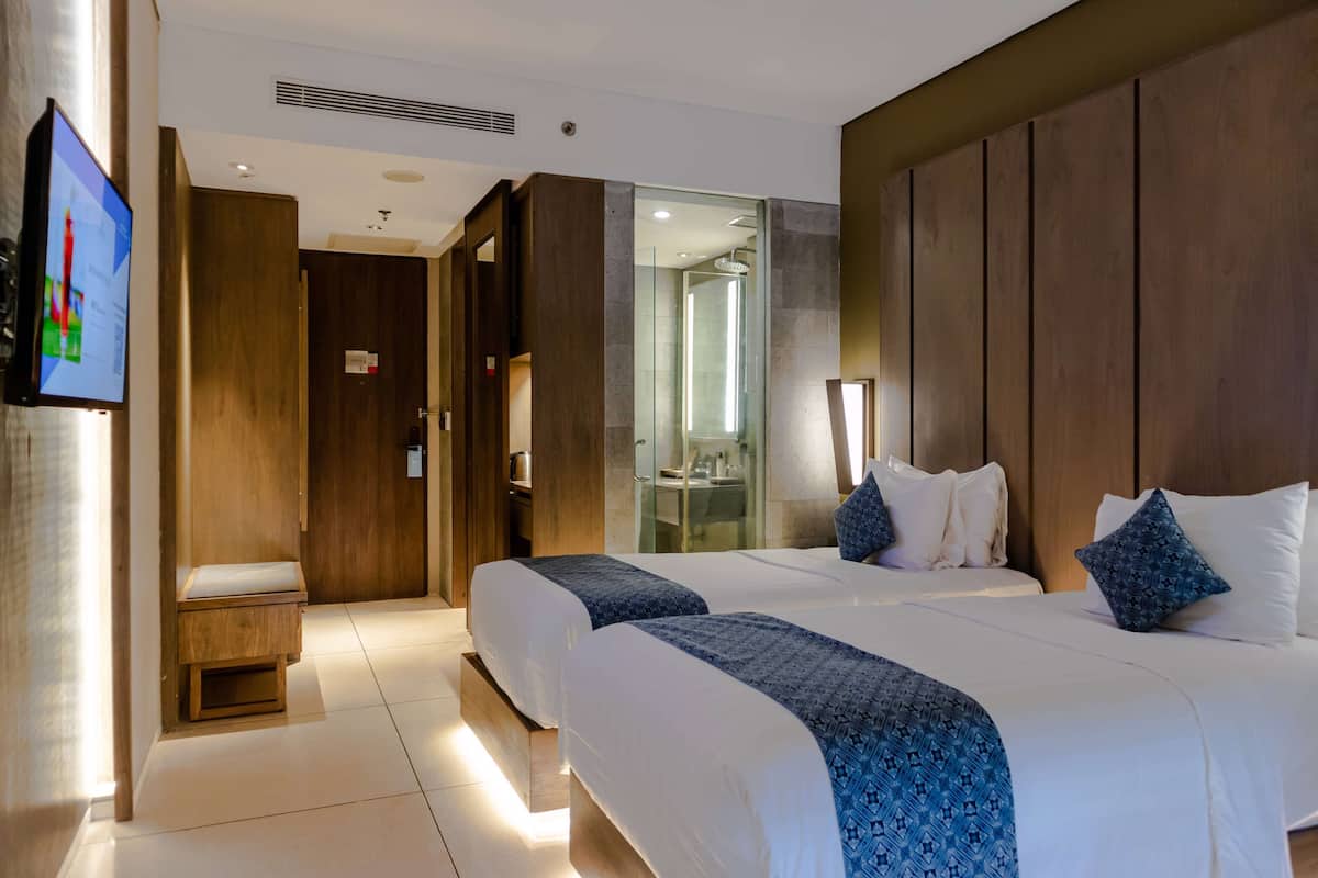 Une chambre d’hôtel avec deux lits, une télévision, un petit coin salon et une salle de bain visible à travers une porte en verre.