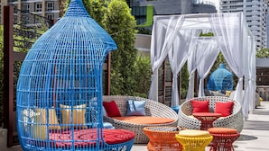 Een dakterras met rieten meubilair, waaronder een blauwe hangstoel, rieten banken en kleine ronde tafels.