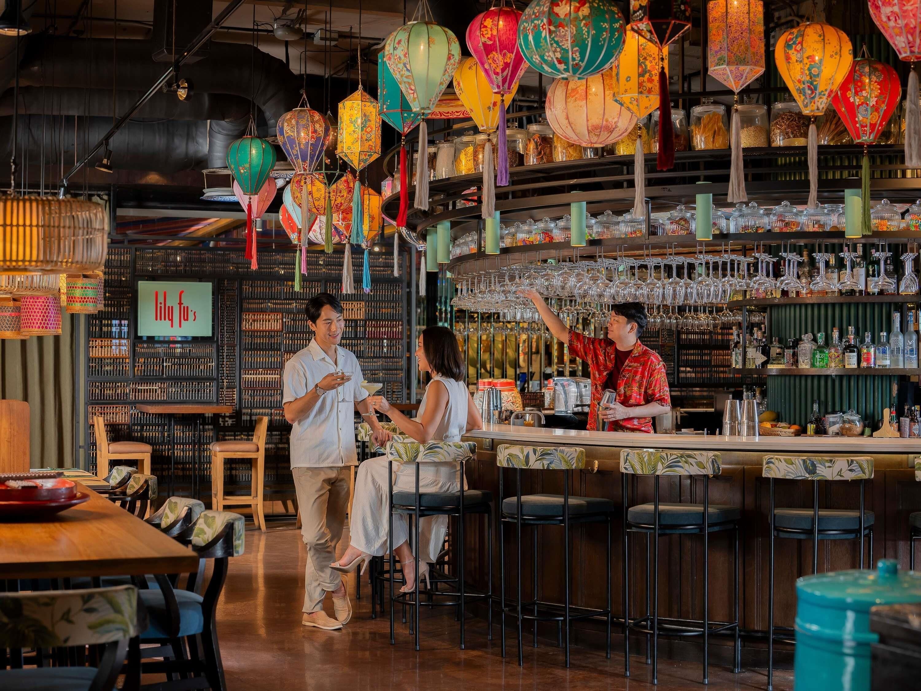 A bar area with a bartender, a customer, and a server. There are colorful lanterns hanging from the ceiling, a bar counter with glasses and bottles, and a seating area with chairs.