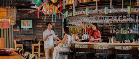 A bar area with a bartender, a customer, and a server. There are colorful lanterns hanging from the ceiling, a bar counter with glasses and bottles, and a seating area with chairs.