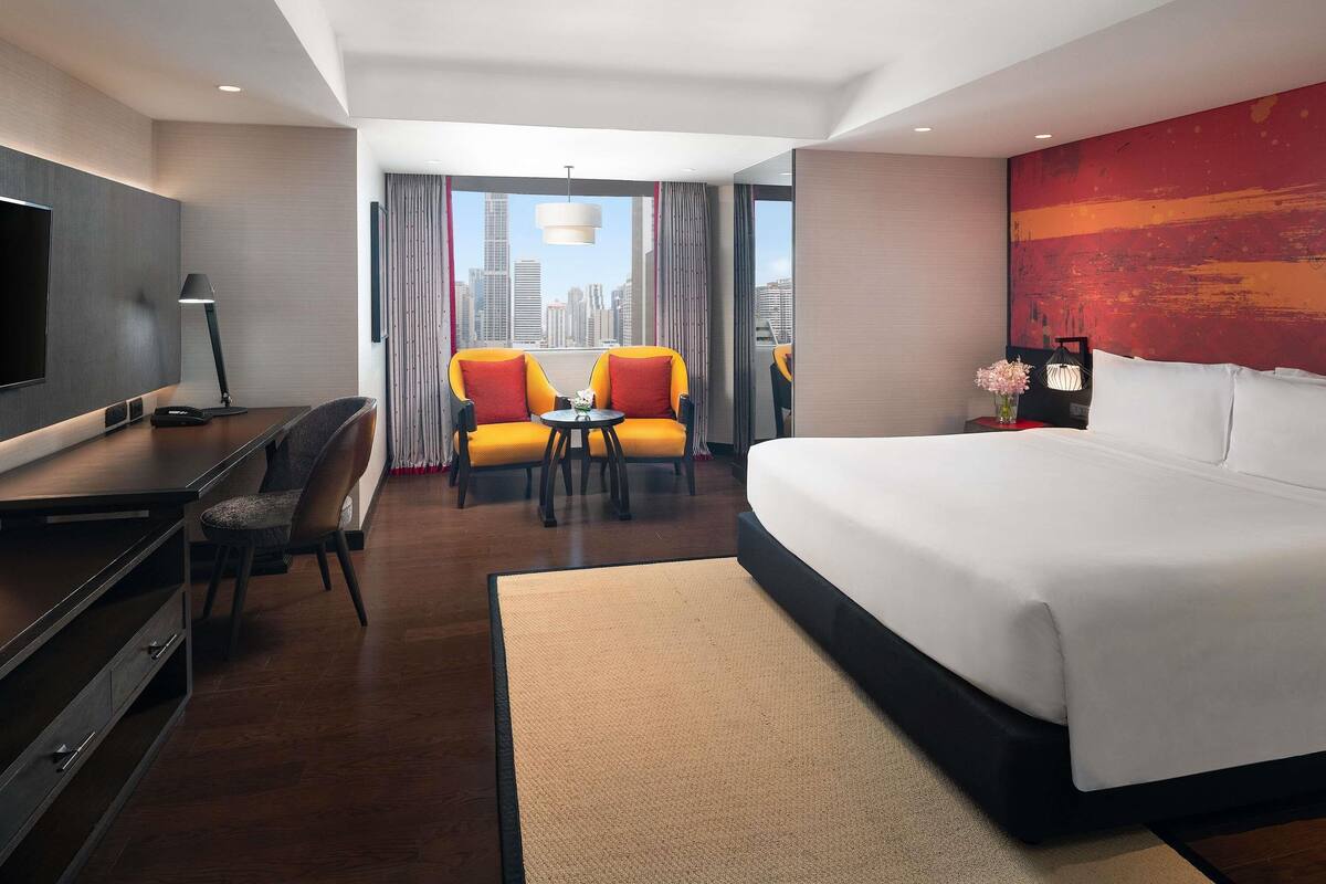 Habitación de hotel con una cama grande, un escritorio con silla, dos sillones, una mesita, televisión y vista al horizonte de la ciudad a través de amplias ventanas.