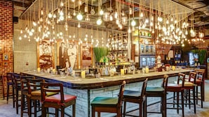 Een bar met meerdere hangende lampen, houten barstoelen en een verscheidenheid aan flessen en glazen op de toog.