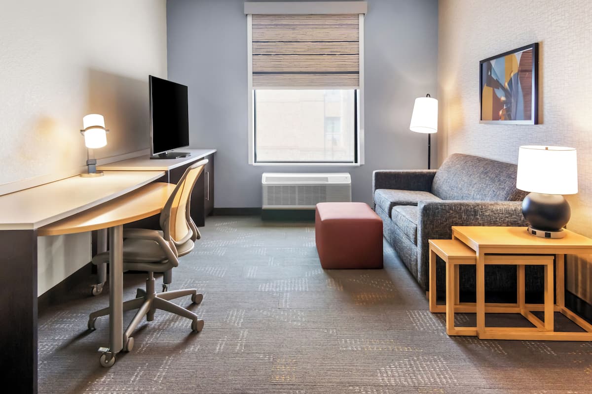 A hotel room with a desk, chair, TV, sofa, ottoman, and two bedside tables with lamps.