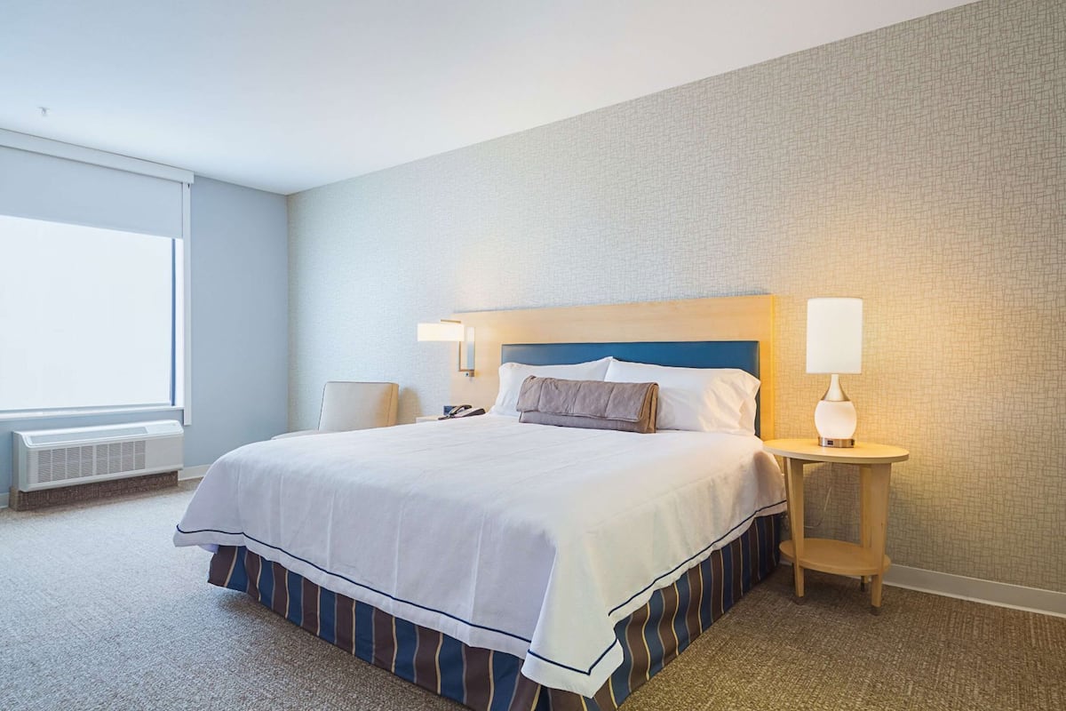 A hotel room with a large bed, a bedside table with a lamp, a window, and a wall heater.