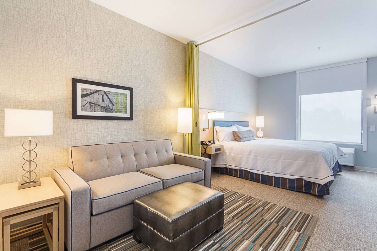 A hotel room with a sofa, a bed, a nightstand, a lamp, a picture on the wall, and a window.