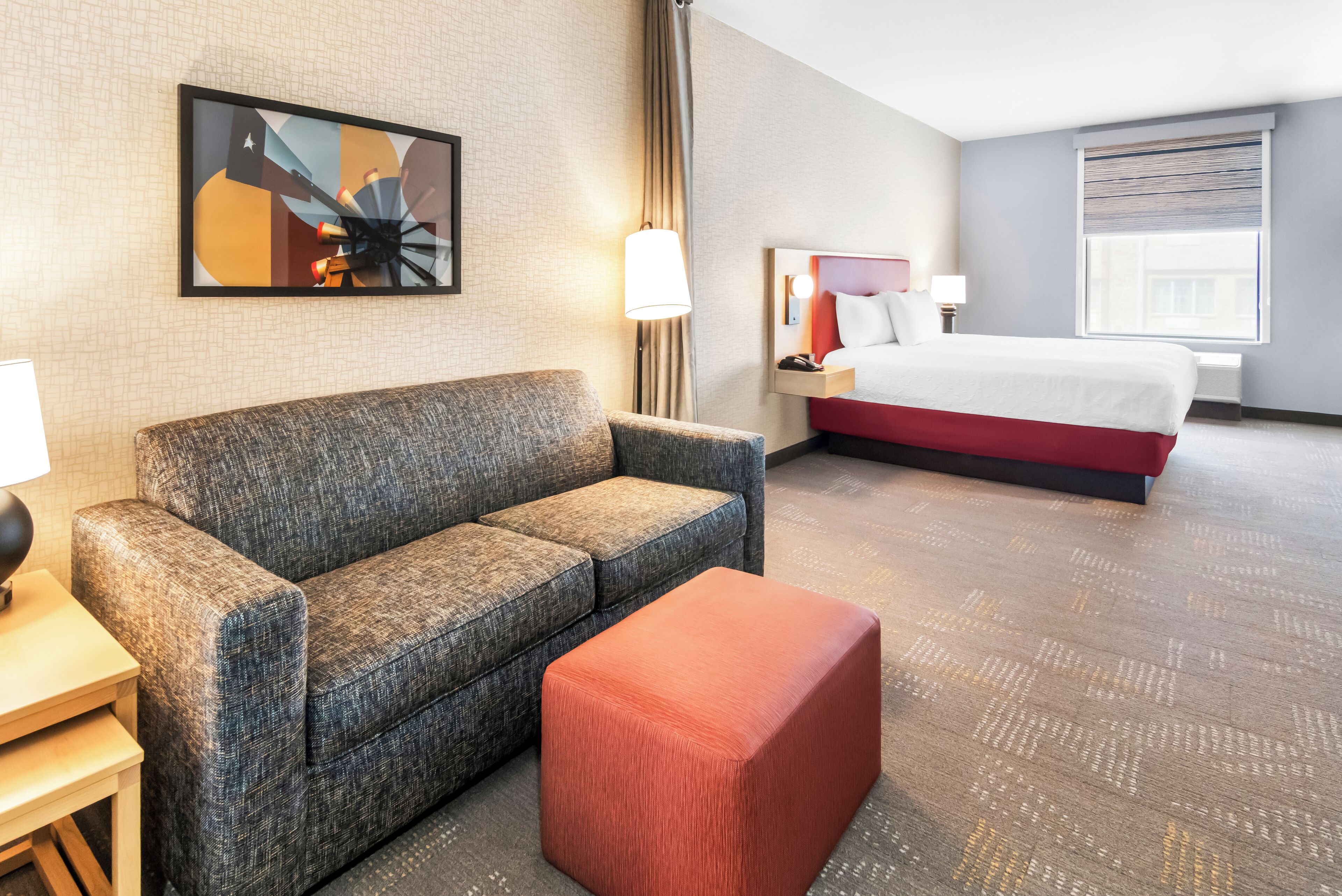 Habitación de hotel con un sofá gris, un reposapiés rojo, una cama con ropa de cama blanca, una mesita de noche con lámpara y un cuadro en la pared.
