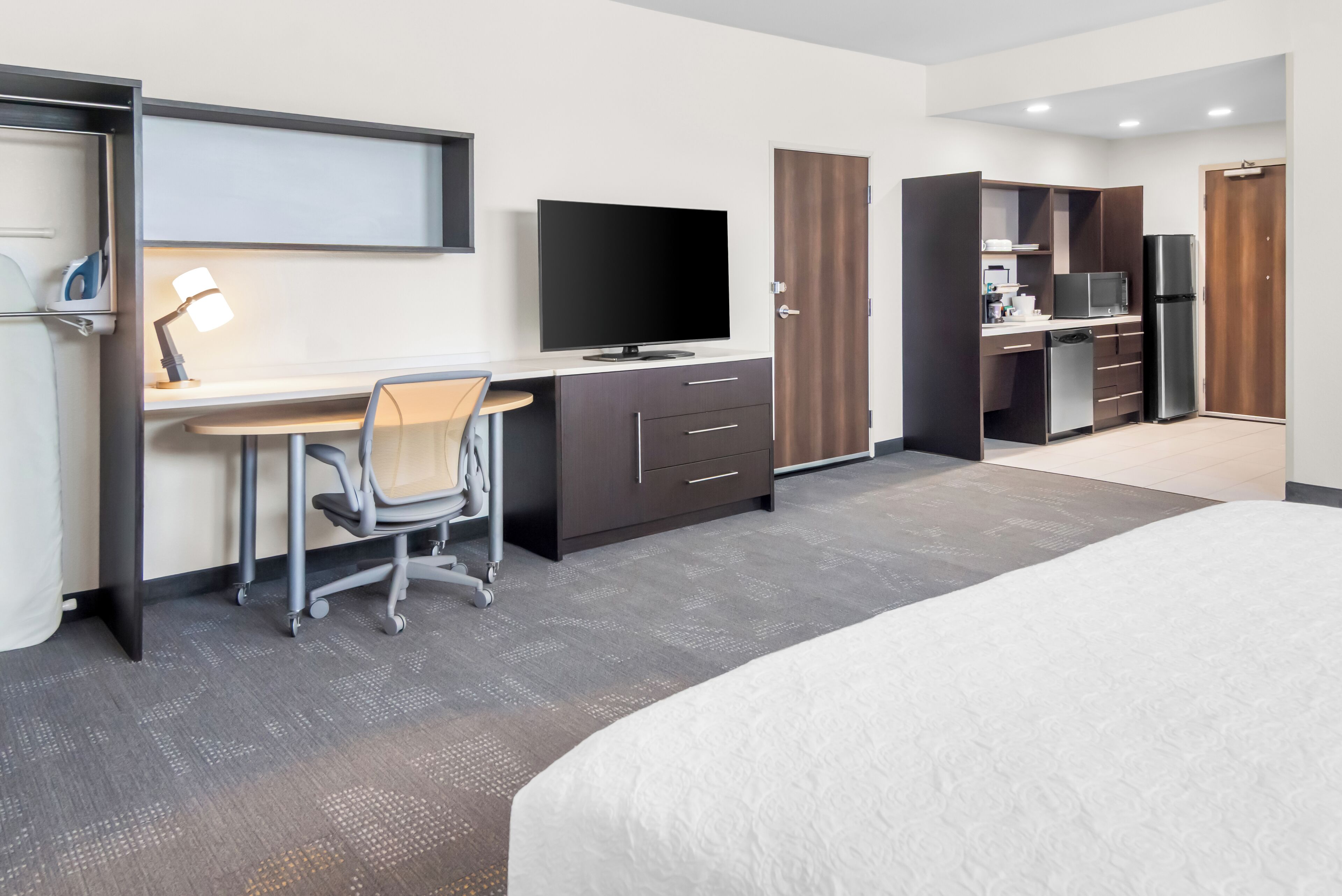 A hotel room with a bed, a desk with a chair, a television, a refrigerator, and a wardrobe.