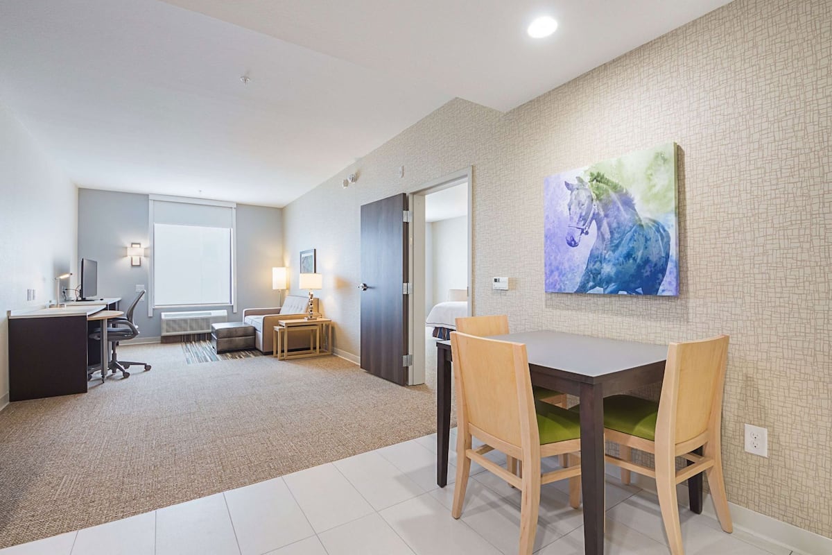 A hotel room with a dining area, a desk with a computer, a sofa, a small table with a lamp, and a large abstract painting on the wall.