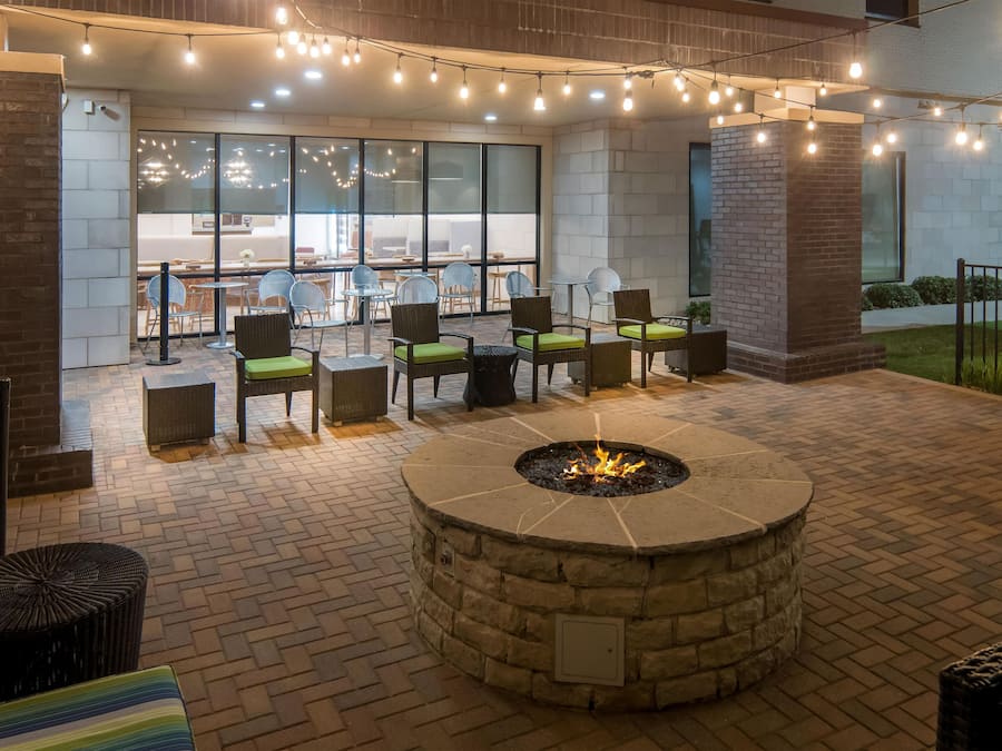 An outdoor seating area with a stone fire pit, surrounded by wicker chairs and tables, under string lights.