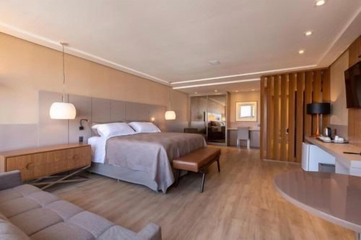 Una habitación de hotel moderna con cama, mesita de noche, banco y una pequeña zona de kitchenette.