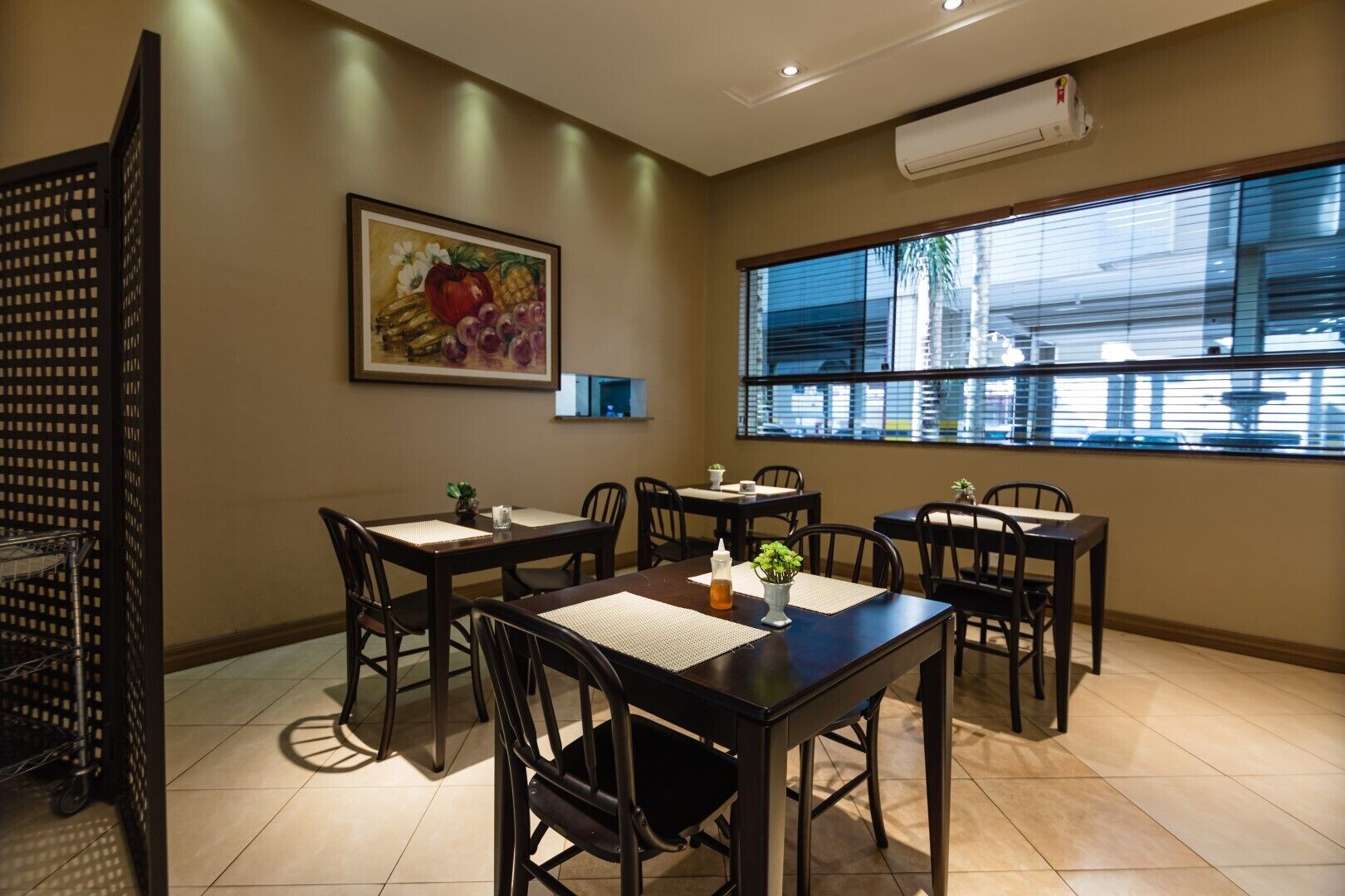 Un espace repas comprenant six tables et chaises, une grande fenêtre avec des stores, un appareil de climatisation et un tableau accroché au mur.